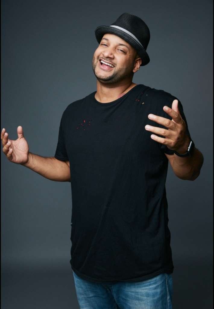 Vibrant man in black t-shirt and fedora smiling and posing confidently for the camera.