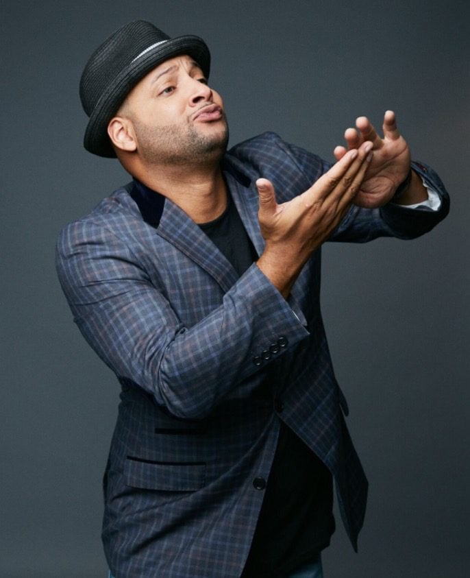 Dapper man in plaid blazer making a gesture with his hands against a dark background.
